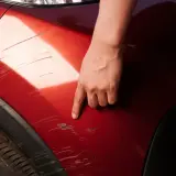 Dłoń wskazuje na porysowane auto. Widać liczne zadrapania na czerwonym lakierze, świadczące o uszkodzeniach.