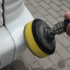 Dłoń trzyma polerkę z żółtą gąbką, która dotyka białego zderzaka. To pokazuje, jak polerować auto, by nadać mu blask.