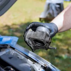Ręka w czarnej rękawiczce trzyma przepustnicę. Zanieczyszczona przepustnica może powodować problemy z silnikiem, takie jak nierówna praca.