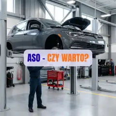 Mechanik pracuje pod samochodem na podnośniku w warsztacie. Napis "ASO - CZY WARTO?" sugeruje dyskusję o serwisach autoryzowanych.