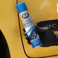 Ręka w czarnej rękawiczce trzyma puszkę K2 Tar Remover, preparatu co rozpuszcza smołę i asfalt z żółtego lakieru samochodu.