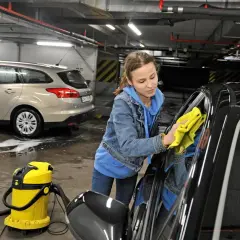 Dziewczyna z żółtą ściereczką poleruje czarne auto. Obok stoi żółty odkurzacz i wiadro. W tle mężczyzna myje srebrne auto. Pokazują, czym umyć auto domowe sposoby.