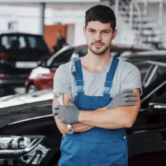 Mechanik w niebieskim kombinezonie, gotowy do wymiany filtrów gazu w samochodzie. W tle inne auta.