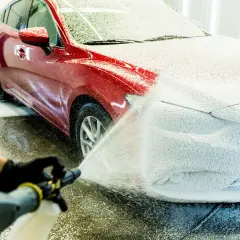 Ręka w czarnej rękawiczce aplikuje mycie aktywną pianą na czerwony samochód. Piana pokrywa maskę i część błotnika.