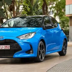 Błękitny Toyota Yaris z czarnym dachem, zaparkowany na podjeździe. W tle budynek i zieleń.