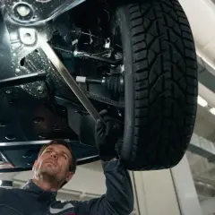 Mechanik bada podwozie auta, szukając przyczyny stukania w kole. Używa latarki, by dokładnie obejrzeć elementy zawieszenia.