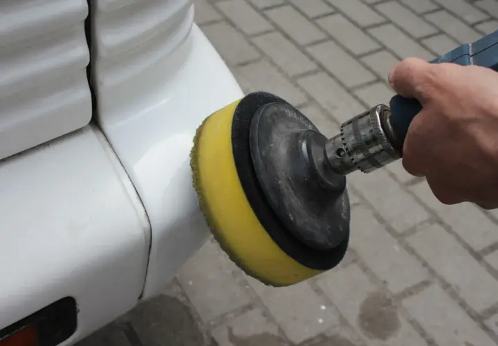 Dłoń trzyma polerkę z żółtą gąbką, która dotyka białego zderzaka. To pokazuje, jak polerować auto, by nadać mu blask.
