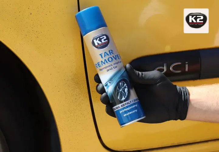 Ręka w czarnej rękawiczce trzyma puszkę K2 Tar Remover, preparatu co rozpuszcza smołę i asfalt z żółtego lakieru samochodu.
