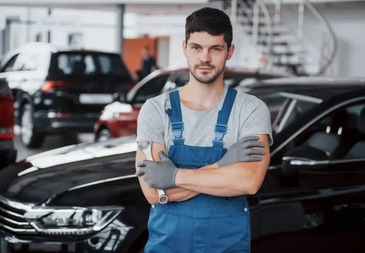 Mechanik w niebieskim kombinezonie, gotowy do wymiany filtrów gazu w samochodzie. W tle inne auta.