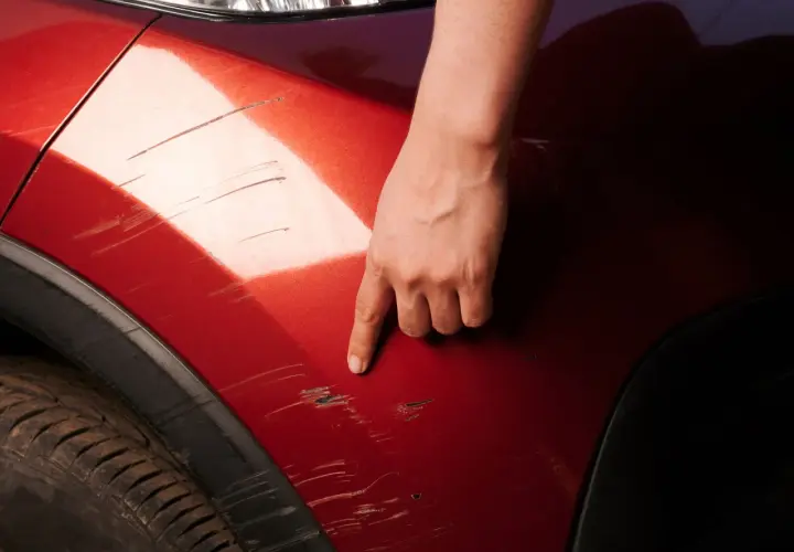 Dłoń wskazuje na porysowane auto. Widać liczne zadrapania na czerwonym lakierze, świadczące o uszkodzeniach.