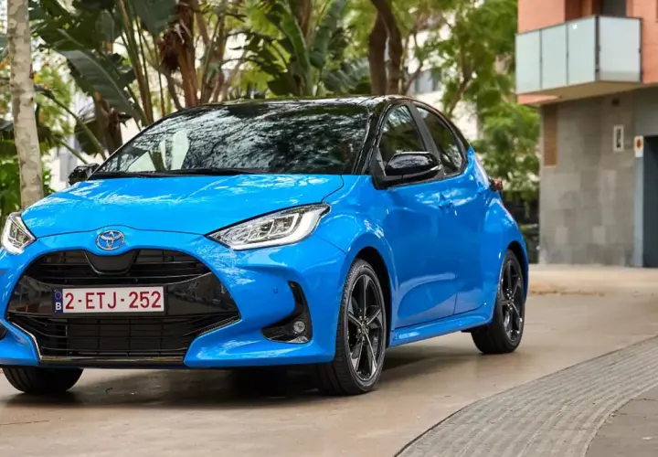 Błękitny Toyota Yaris z czarnym dachem, zaparkowany na podjeździe. W tle budynek i zieleń.