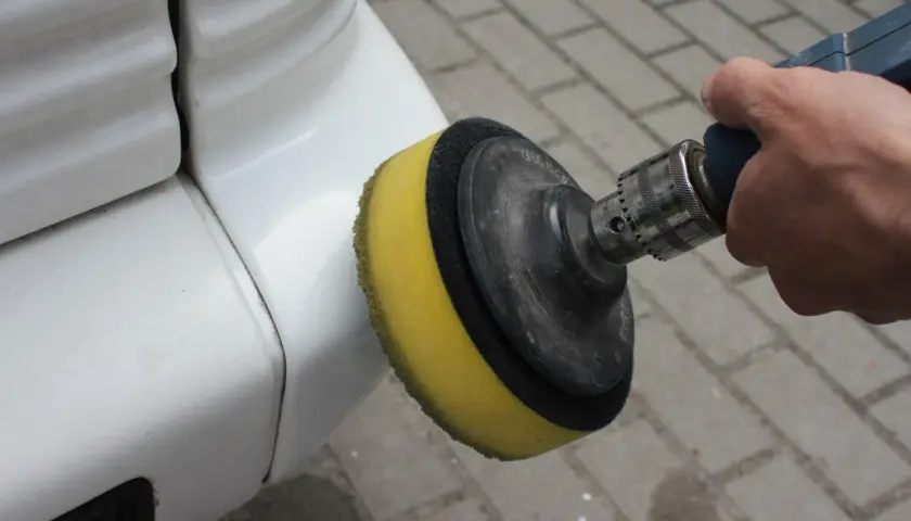 Dłoń trzyma polerkę z żółtą gąbką, która dotyka białego zderzaka. To pokazuje, jak polerować auto, by nadać mu blask.