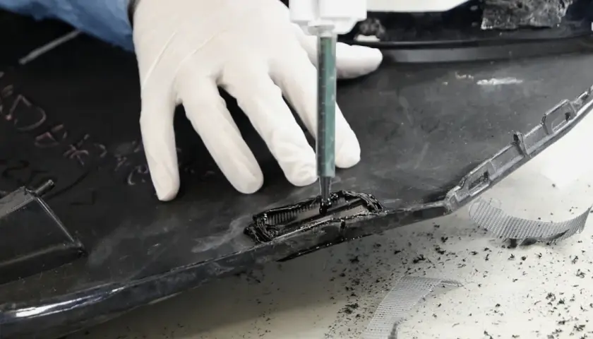 Rękawiczki i strzykawka pomagają w procesie, jak naprawić pęknięty plastik. Widać czarne tworzywo i siatkę wzmacniającą.