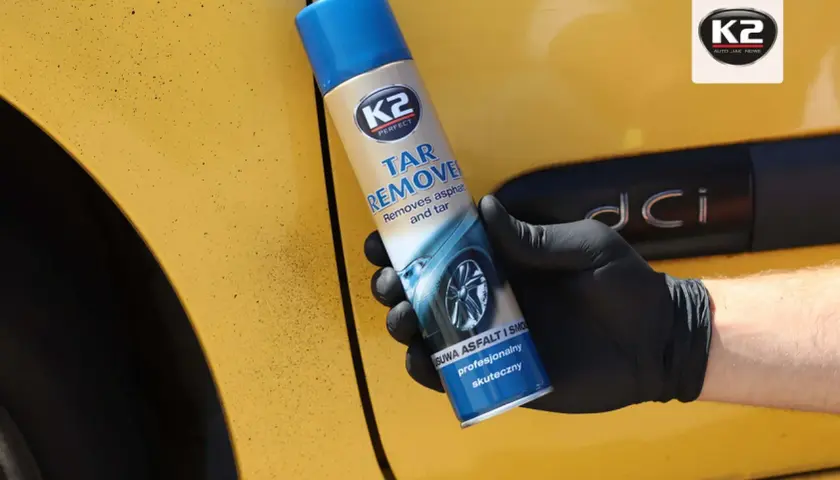 Ręka w czarnej rękawiczce trzyma puszkę K2 Tar Remover, preparatu co rozpuszcza smołę i asfalt z żółtego lakieru samochodu.