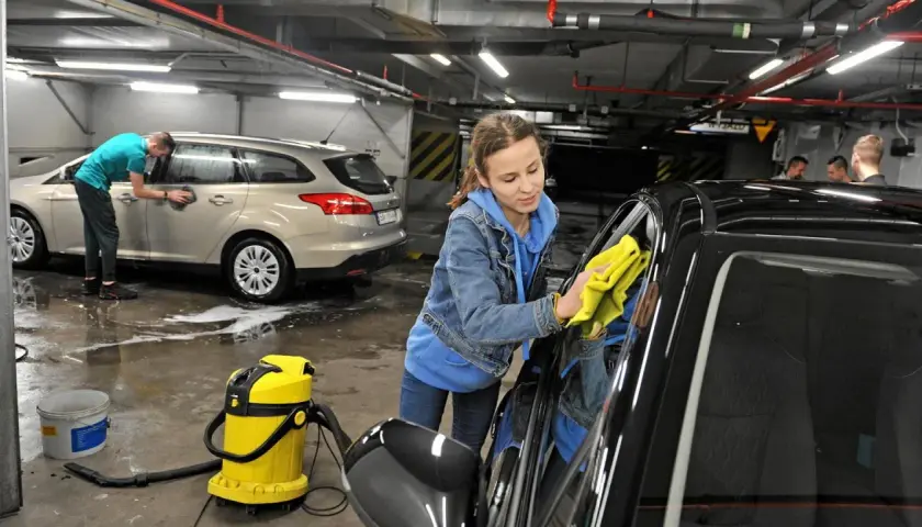 Dziewczyna z żółtą ściereczką poleruje czarne auto. Obok stoi żółty odkurzacz i wiadro. W tle mężczyzna myje srebrne auto. Pokazują, czym umyć auto domowe sposoby.