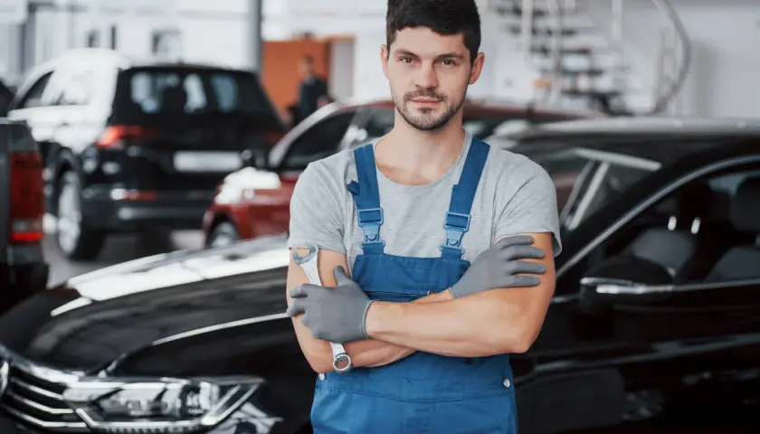 Mechanik w niebieskim kombinezonie, gotowy do wymiany filtrów gazu w samochodzie. W tle inne auta.
