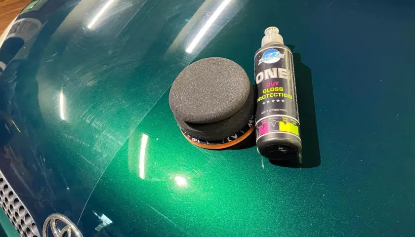 Zestaw do ręcznego polerowania auta: gąbka i butelka z preparatem "ONE CUT GLOSS PROTECTION". Widać zarysowania na zielonym lakierze.