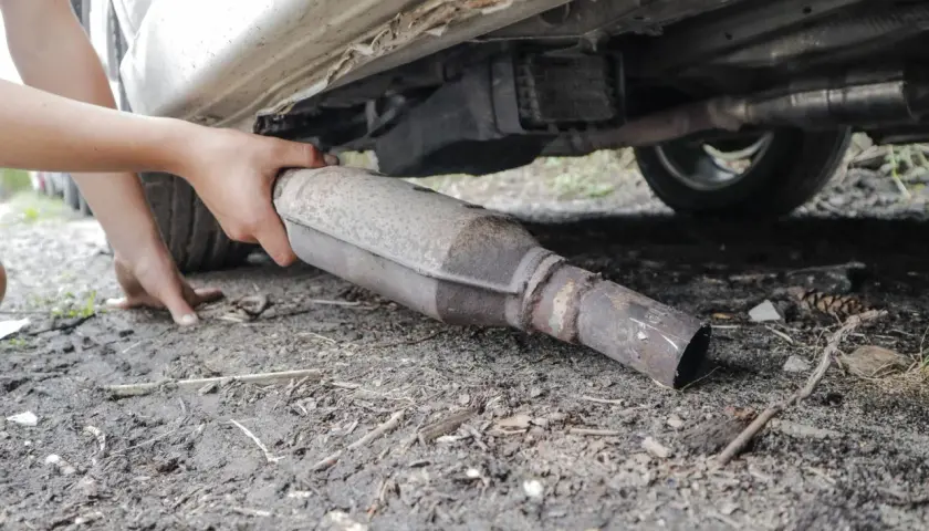 Ręce wyciągają z podwozia samochodu skorodowany element układu wydechowego, prawdopodobnie po wycinaniu katalizatora.