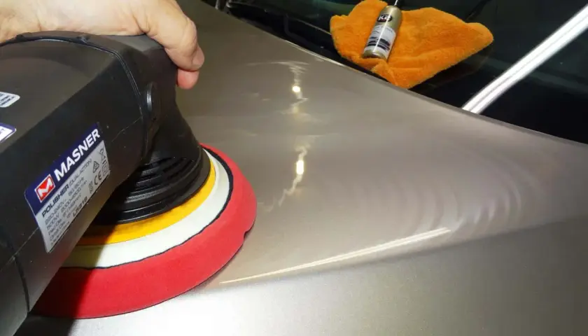 Nakładanie powłoki ceramicznej na lakier samochodu za pomocą polerki. Pomarańczowa ściereczka i buteleczka z preparatem K2 na masce.