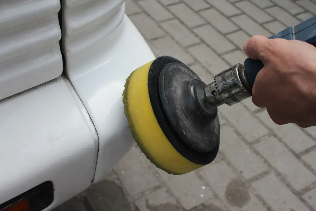 Dłoń trzyma polerkę z żółtą gąbką, która dotyka białego zderzaka. To pokazuje, jak polerować auto, by nadać mu blask.