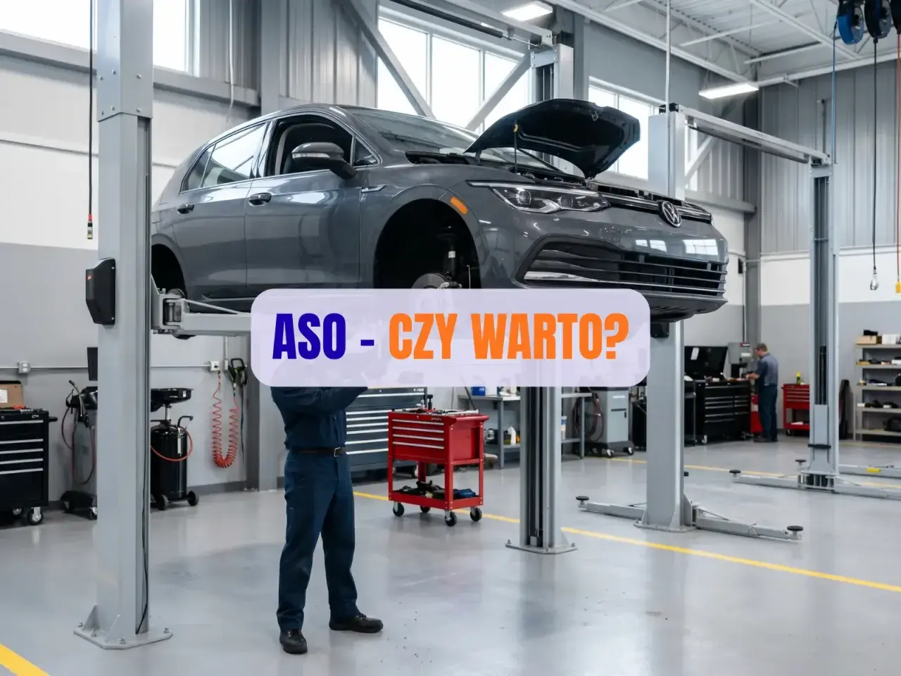 Mechanik pracuje pod samochodem na podnośniku w warsztacie. Napis "ASO - CZY WARTO?" sugeruje dyskusję o serwisach autoryzowanych.