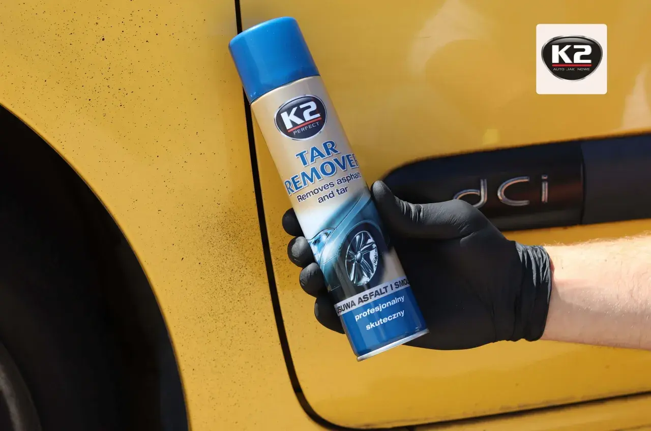 Ręka w czarnej rękawiczce trzyma puszkę K2 Tar Remover, preparatu co rozpuszcza smołę i asfalt z żółtego lakieru samochodu.