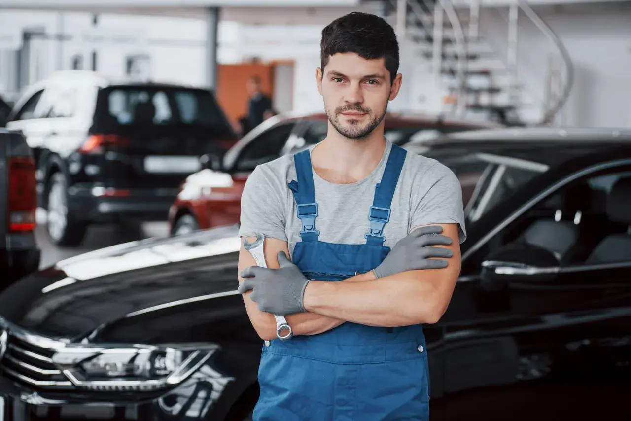 Mechanik w niebieskim kombinezonie, gotowy do wymiany filtrów gazu w samochodzie. W tle inne auta.