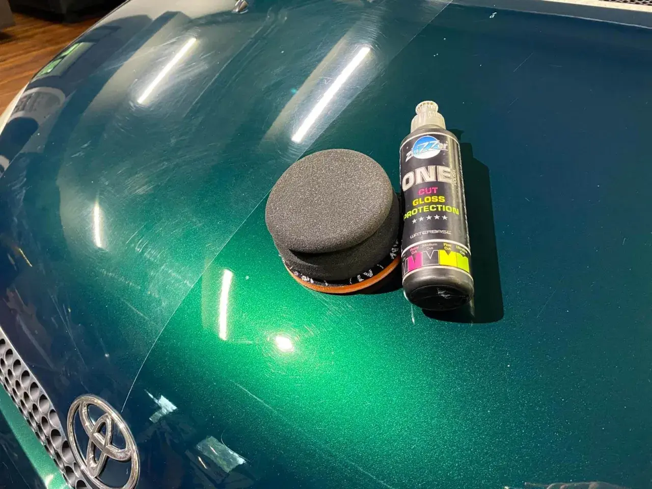 Zestaw do ręcznego polerowania auta: gąbka i butelka z preparatem "ONE CUT GLOSS PROTECTION". Widać zarysowania na zielonym lakierze.