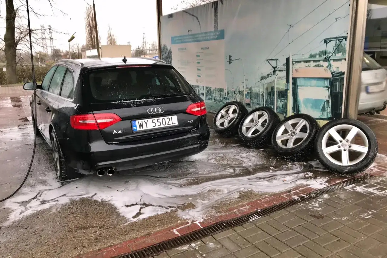 Czarny Audi A4 na myjni bezdotykowej, obok cztery felgi. Poznaj myjnia bezdotykowa triki, by Twoje auto lśniło!
