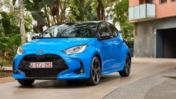 Błękitny Toyota Yaris z czarnym dachem, zaparkowany na podjeździe. W tle budynek i zieleń.